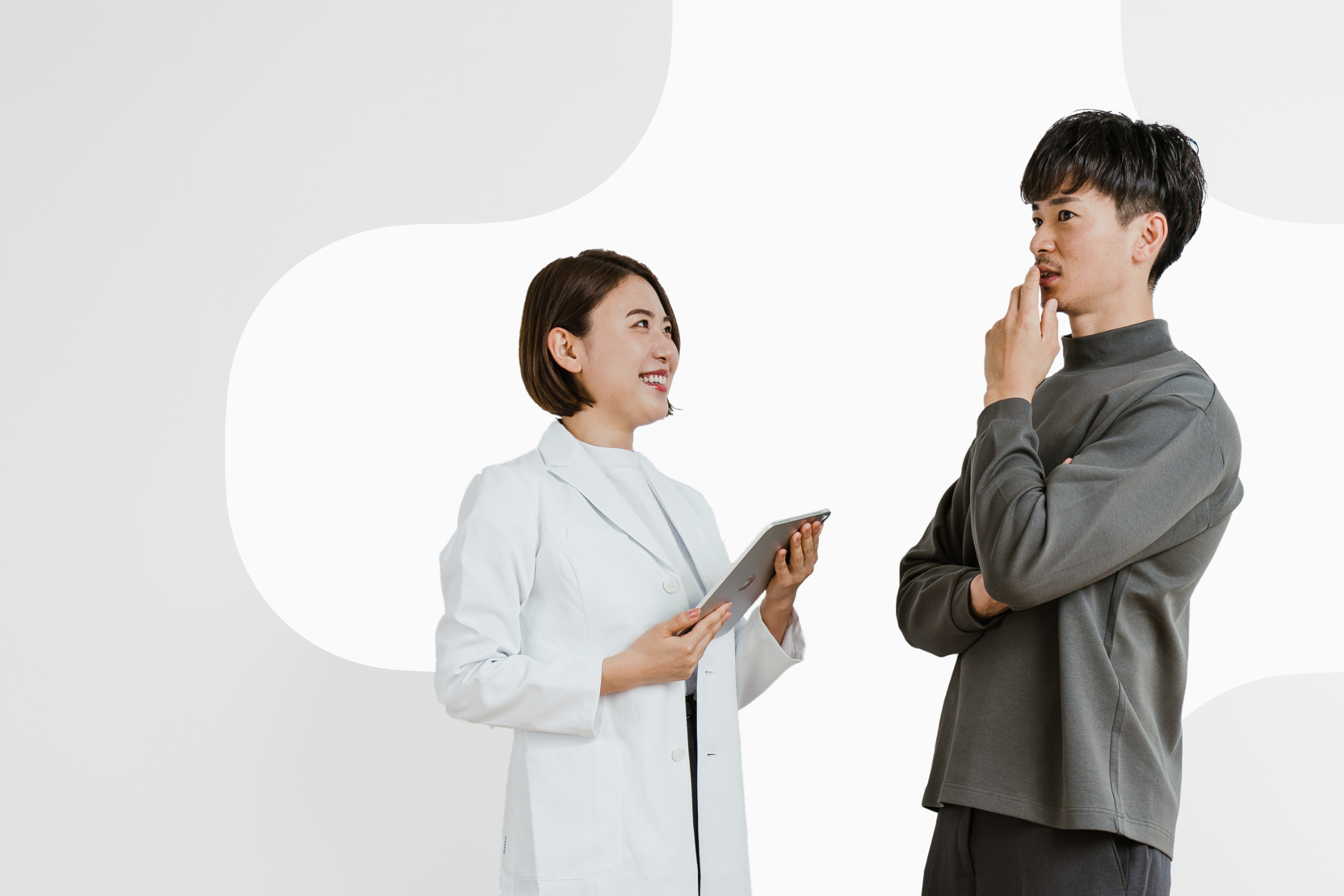 画像は2人の若い男女を描いています。女性は白衣を着て微笑んでいます。男性は灰色のシャツを着て、考え込むようなポーズをとりながら女性の方を見ています。2人は何かを議論したり考えたりしているようです。背景は白一色で、人物だけが際立っています。プロフェッショナルな雰囲気の中にも、2人の自然体な表情から親しみやすさも感じられる1枚です。