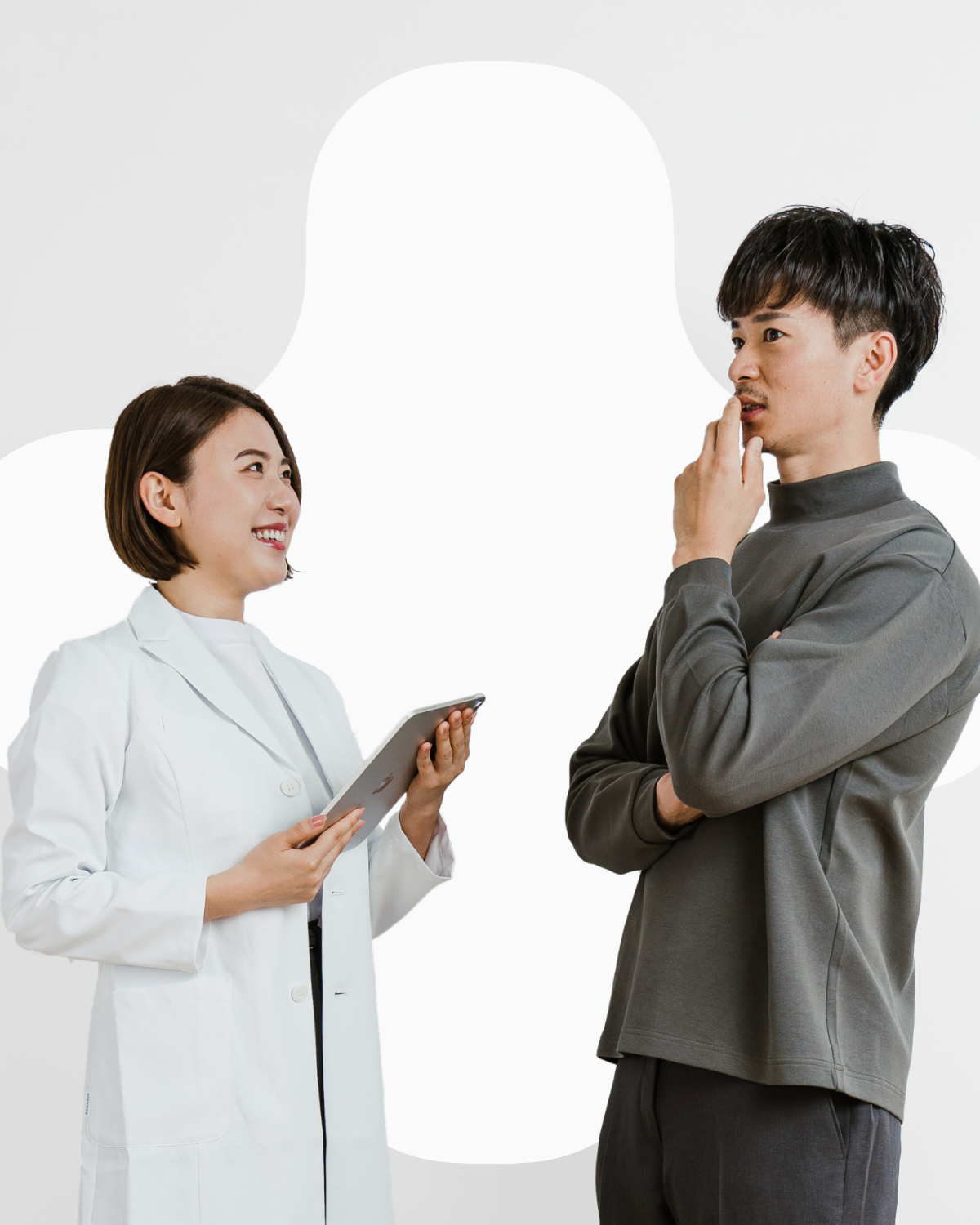 画像は2人の若い男女を描いています。女性は白衣を着て微笑んでいます。男性は灰色のシャツを着て、考え込むようなポーズをとりながら女性の方を見ています。2人は何かを議論したり考えたりしているようです。背景は白一色で、人物だけが際立っています。プロフェッショナルな雰囲気の中にも、2人の自然体な表情から親しみやすさも感じられる1枚です。